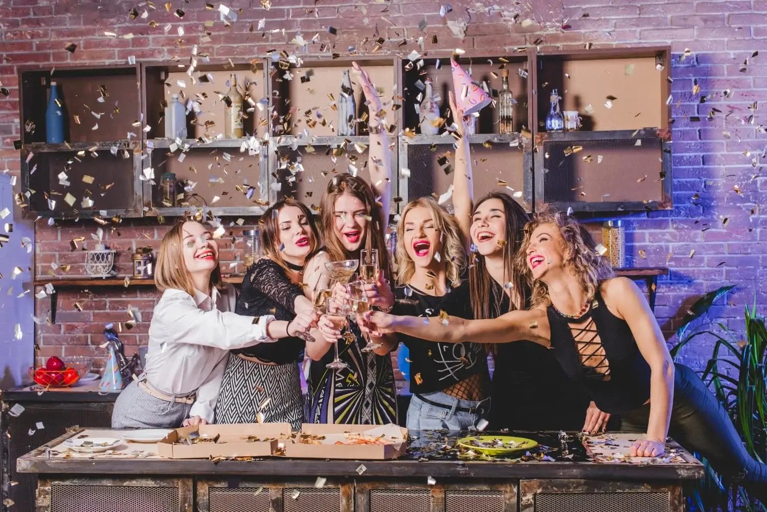 Six women celebrating indoors with glasses and confetti, joyful scene.