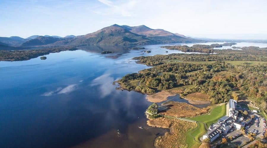 Aerial lake view surrounded by forest and mountains under blue sky.